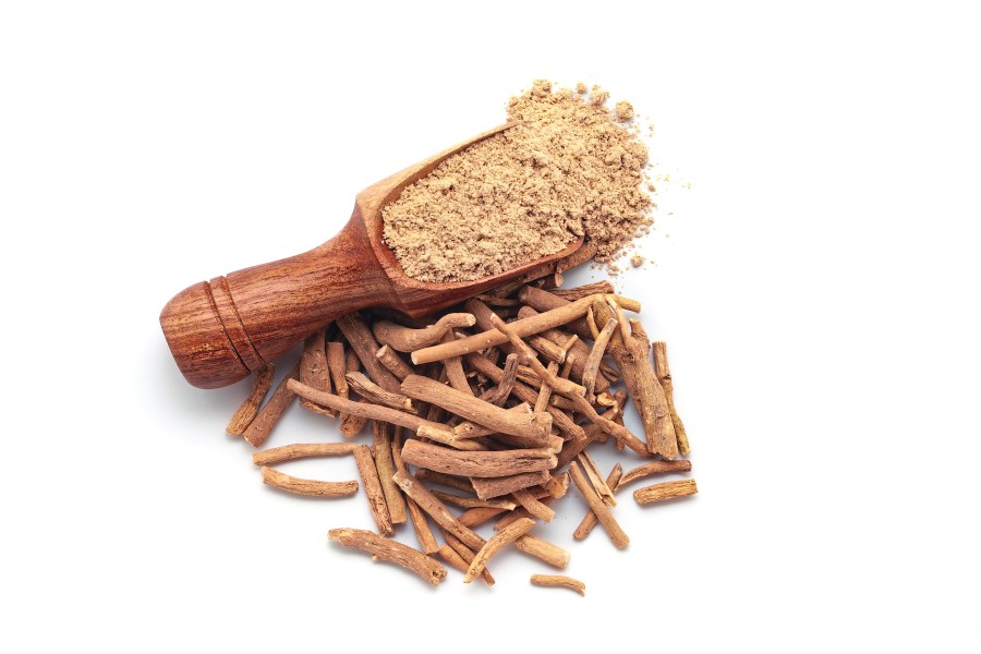 A wooden scoop filled with organic Ashwagandha (Withania somnifera) powder, placed beside a heap of ashwagandha roots, isolated on a white background.