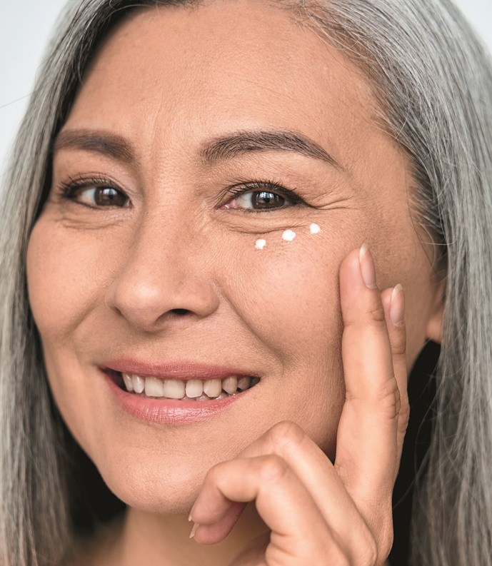 Happy middle aged mature asian woman, senior older 50 year lady looking at camera touching her face applying eye antiaging anti wrinkle skin care cream, isolated on white close up face portrait.