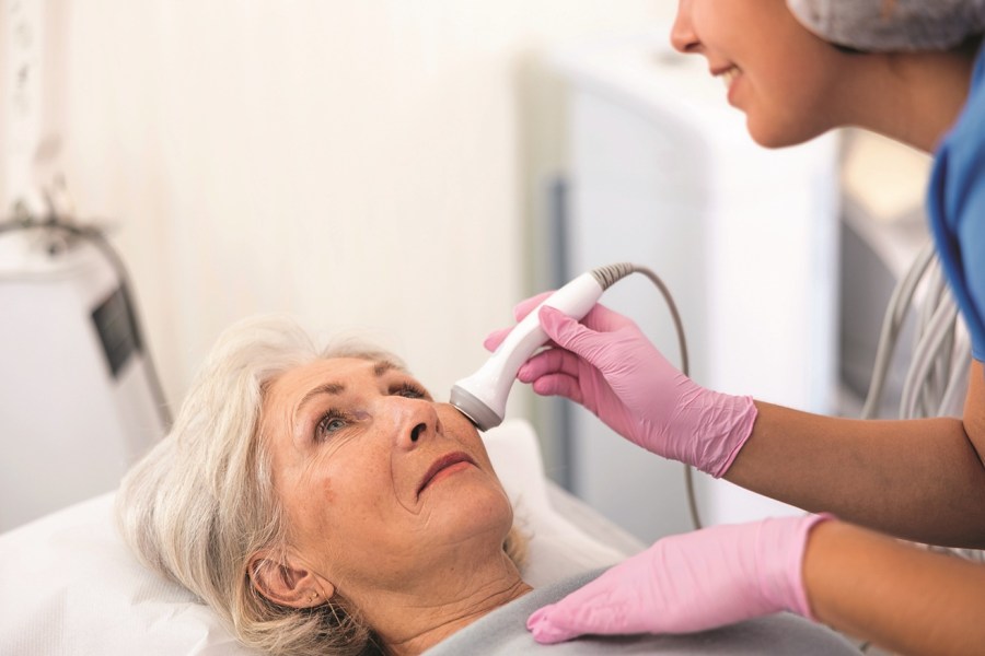 Professional young female cosmetologist doing facial skin ultrasound cavitation for for face lifting of mature woman laying in beauty salon